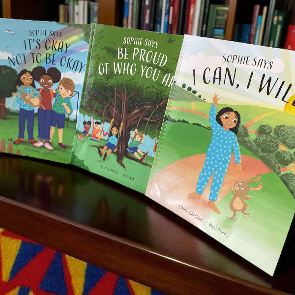 Three children's books titled 'It's Okay Not to Be Okay', 'Be Proud of Who You Are', and 'I Can, I Will' on a shelf with a colorful rug underneath.