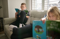 Two children sitting on a couch, one reading a book and the other looking at a toy box.