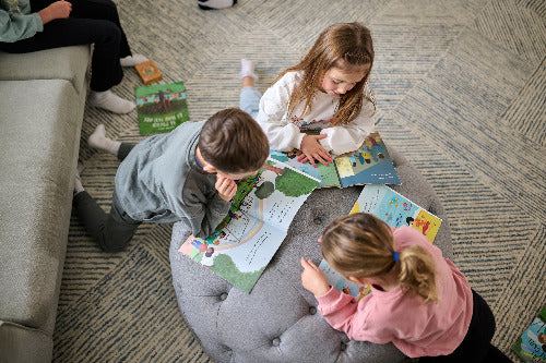 Three children sitting on a gray ottoman reading books on a carpeted floor.
