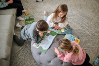 Three children sitting on a gray ottoman reading books on a carpeted floor.
