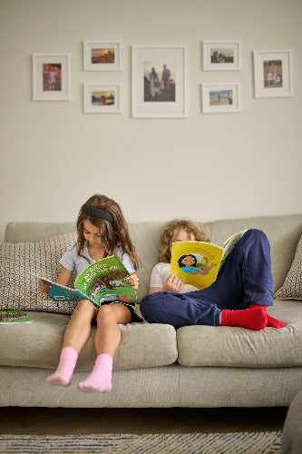 Two children reading books on a couch with framed pictures on the wall.
