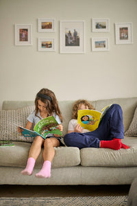 Two children reading books on a couch with framed pictures on the wall.