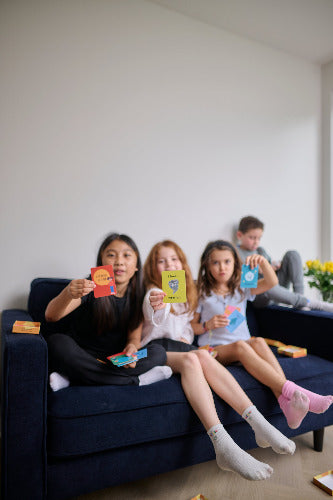 Four children sitting on a couch holding colorful cards in a room with a white wall and a plant.