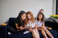 Three children sitting on a couch playing with a device, with another person partially visible in the background.