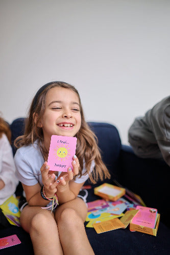Young girl holding a pink card with 'I feel happy' text, sitting on a couch with colorful cards around.