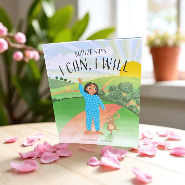 Children's book titled 'I Can, I Will' by Sophie Murphy on a table with pink petals and plants in the background.